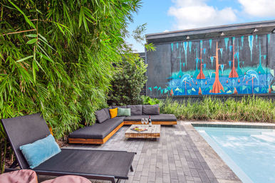 Chill modern poolside patio with L-shaped black sectional, bamboo privacy hedge, colorful mural of stylized trees and fish, low wooden coffee table with wine and glasses, and lounge chairs beside a swimming pool.