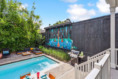 Sunny modern backyard patio with a small rectangular pool, vibrant tropical mural on a dark privacy wall, tall bamboo screening, low sectional lounge with colorful cushions, outdoor dining table and stainless steel grill.