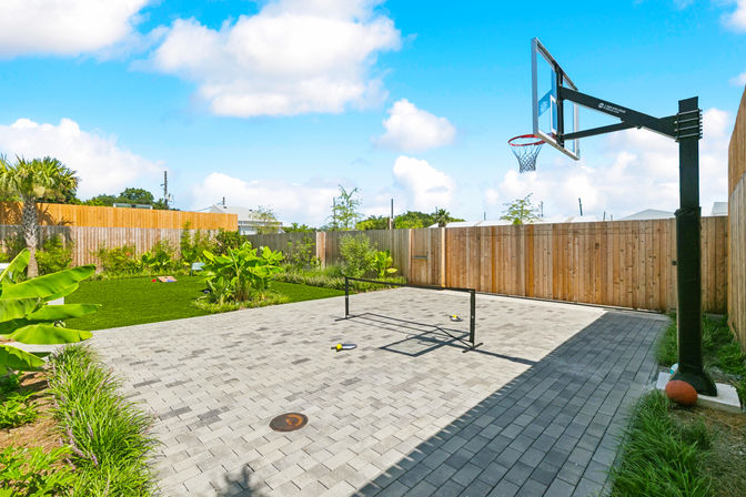 Sunny suburban backyard with gray brick paved court, adjustable basketball hoop, portable pickleball net, green lawn, tropical plants and wooden privacy fence under a blue sky