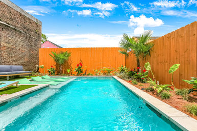 Sunny backyard swimming pool with turquoise water, wooden privacy fence, tropical plants and cozy lounge seating beside a brick wall — bright, inviting oasis.