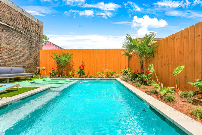 Sunny backyard swimming pool with turquoise water, wooden privacy fence, tropical plants and cozy lounge seating beside a brick wall — bright, inviting oasis.
