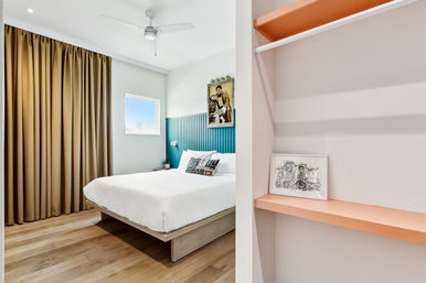 Sunlit modern minimalist apartment bedroom with teal paneled headboard, white bedding, wood floors, tan curtains, ceiling fan and open closet shelf with framed sketch.