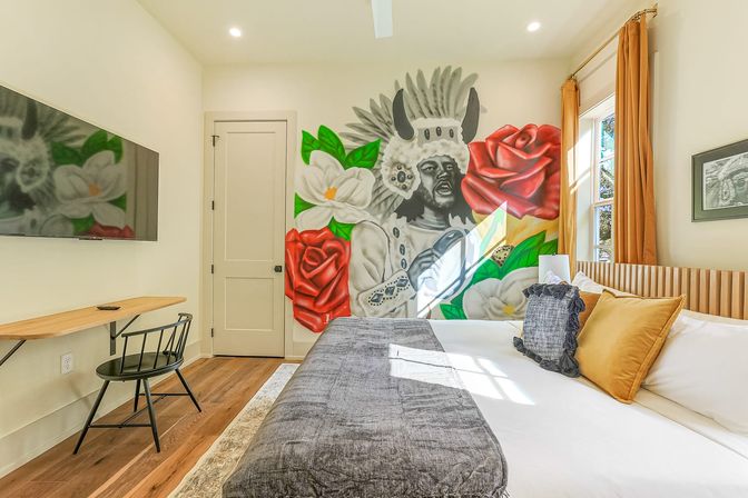 Sunny bedroom with large bed and hardwood floors, wall-mounted TV and desk, featuring a bold mural of a person in a feathered headdress surrounded by red roses and white magnolia flowers.
