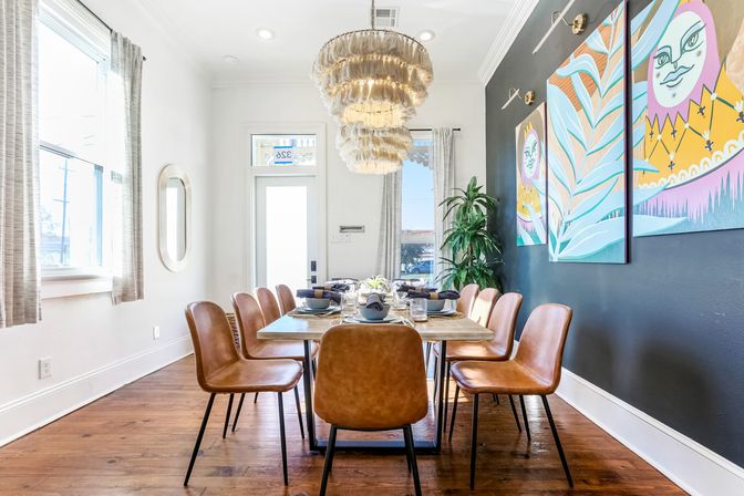 Sunlit modern dining room with a long wooden table set for eight, tan leather chairs, a layered fringe chandelier, colorful artwork on a dark accent wall, tall windows and hardwood floors.