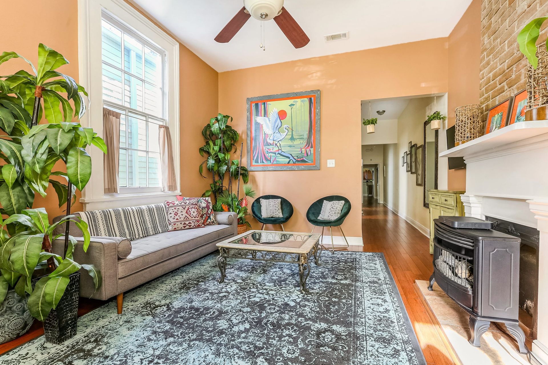 Sunlit urban townhouse living room with warm peach walls, mid-century gray sofa, patterned rug, glass coffee table, lush potted plants, colorful wall art, ceiling fan and small fireplace stove on hardwood floors.