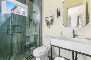 Sunlit modern residential bathroom with deep green tiled walk-in shower and glass door, white toilet, minimalist white sink on a black metal stand, arched mirror and hanging eucalyptus wall decor.