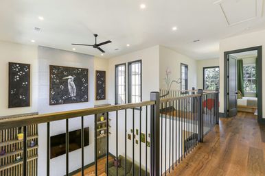 Bright modern second-floor landing in an open-concept home with warm hardwood floors, black metal railing overlooking a living area, oversized framed heron artwork and floral panels, ceiling fan, tall windows, and an open doorway to a bedroom.