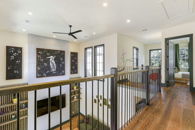 Bright modern second-floor landing in an open-concept home with warm hardwood floors, black metal railing overlooking a living area, oversized framed heron artwork and floral panels, ceiling fan, tall windows, and an open doorway to a bedroom.