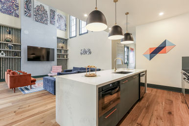 Bright open-concept living room and kitchen with white waterfall island, black pendant lights, hardwood floors, colorful sofas, built-in shelving, flat-screen TV and geometric wall art.