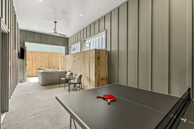 Covered outdoor living space with a black ping-pong table and red paddles in the foreground, hot tub and two chairs behind a wooden privacy partition, wall-mounted TV and ceiling fan on modern board-and-batten walls.