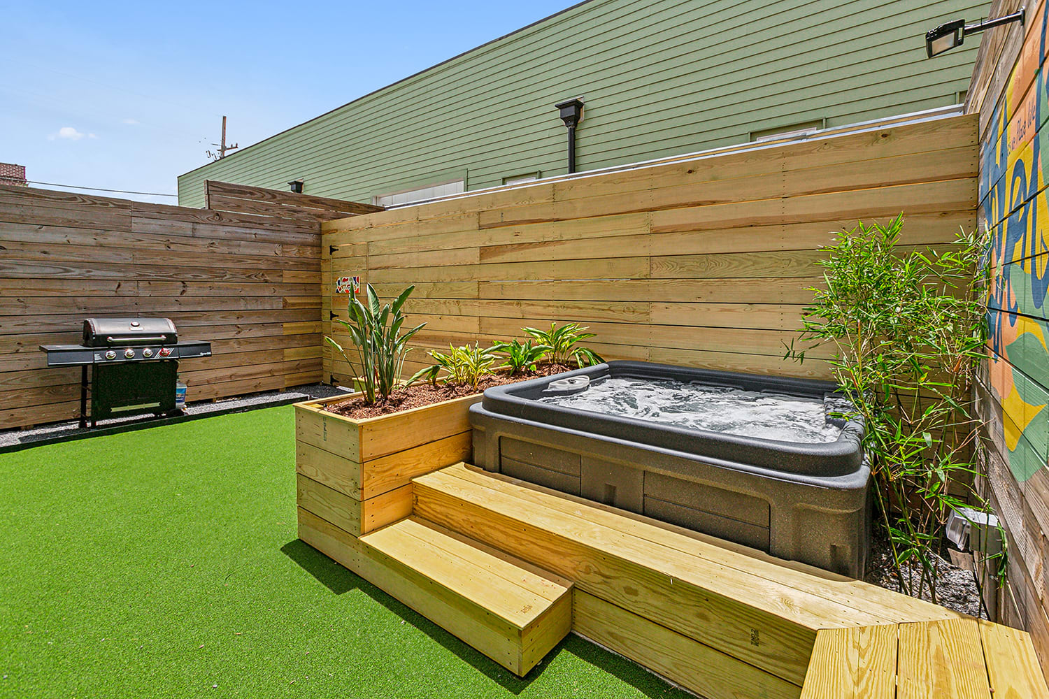 Cozy urban patio with bubbling hot tub set into wooden decking, raised planter of tropical greenery, bright artificial turf, gas grill by a wooden privacy fence and a colorful mural.