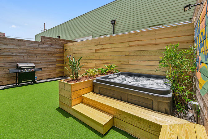 Cozy urban patio with bubbling hot tub set into wooden decking, raised planter of tropical greenery, bright artificial turf, gas grill by a wooden privacy fence and a colorful mural.