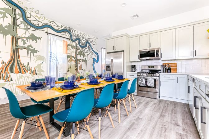Open-plan modern kitchen and dining area with a long wooden table set for eight with blue chairs and dinnerware, stainless steel appliances, white cabinetry, light wood flooring, and a colorful map-style river and tree mural on the wall.