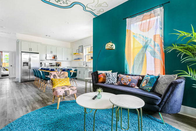 Vibrant open-concept living room and kitchen with a teal accent wall, velvet navy sofa with colorful throw pillows, turquoise shag rug, patterned armchair, round nesting coffee tables, modern dining table with teal chairs and stainless steel refrigerator.