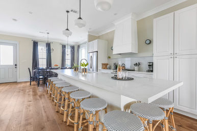 Bright modern farmhouse open-concept kitchen with a long white quartz island lined with woven bistro bar stools, white shaker cabinets, stainless refrigerator, pendant lights, hardwood floors and a sunlit dining nook.