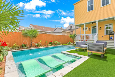 Sunny suburban backyard with turquoise plunge pool featuring built-in submerged loungers and small water fountains, modern gray patio seating on artificial turf beside a yellow two-story house with covered porch, wooden privacy fence and tropical plants under a bright blue sky.