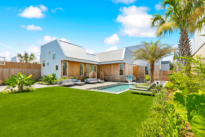 Sunny backyard oasis with a modern coastal-style home (metal roofs, wood siding), rectangular swimming pool with lounge chairs and outdoor sofas, manicured lawn and tropical palms under a bright blue sky.