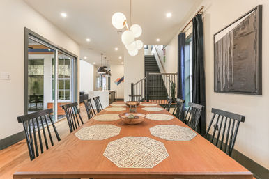 Modern open-concept dining room in a contemporary home: long wooden table with hexagonal woven placemats, black spindle chairs, globe pendant lights, adjacent kitchen island and staircase.