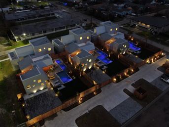 Aerial night view of modern clustered townhouses with private fenced courtyards, glowing blue swimming pools, warm exterior lighting and concrete driveways in an urban neighborhood.