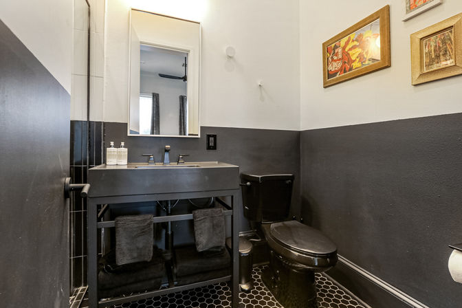 Moody black-and-white powder room with a matte black sink vanity, matching black toilet, hexagon tile floor, rectangular mirror, and framed wall art