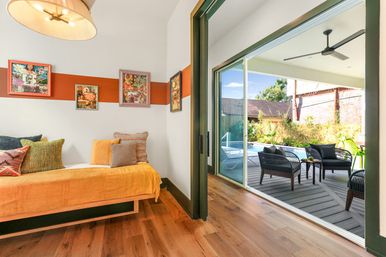 Bright contemporary bedroom with mustard daybed and colorful pillows, hardwood floors and art-accented wall opening through sliding glass doors to a covered poolside deck with wicker lounge chairs, table, ceiling fan and garden