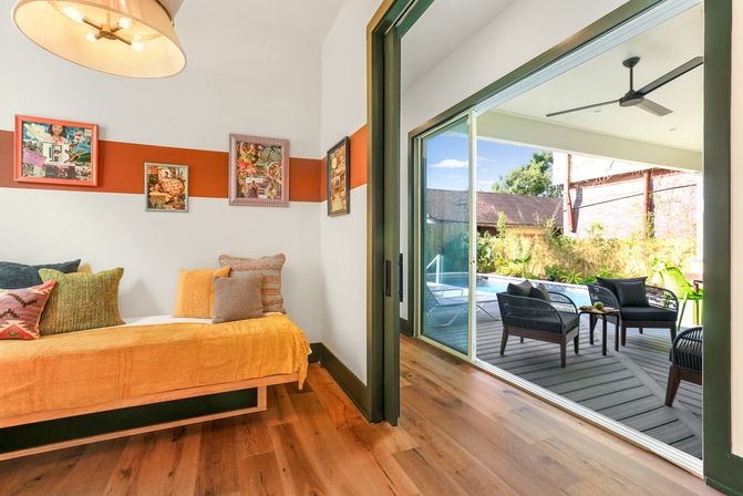 Bright contemporary bedroom with mustard daybed and colorful pillows, hardwood floors and art-accented wall opening through sliding glass doors to a covered poolside deck with wicker lounge chairs, table, ceiling fan and garden