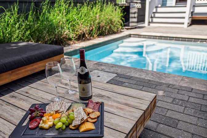 Poolside charcuterie board with assorted cheeses, grapes, crackers and cured meats, plus a bottle of red wine and two wine glasses on a wooden table by a backyard pool and patio lounge.