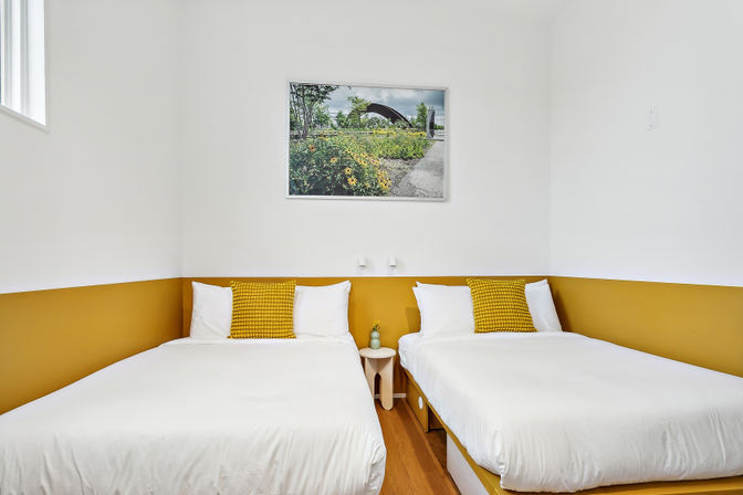 Bright minimalist guest room with two white single beds, mustard-colored accent band and matching throw pillows, small round bedside table with vase, hardwood floor and framed park landscape above