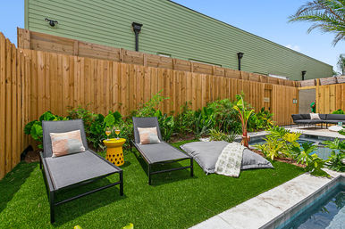 Residential backyard poolside lounge with two gray chaise lounges, yellow side table with drinks, oversized floor cushion on artificial turf, tropical plants and wooden privacy fence.