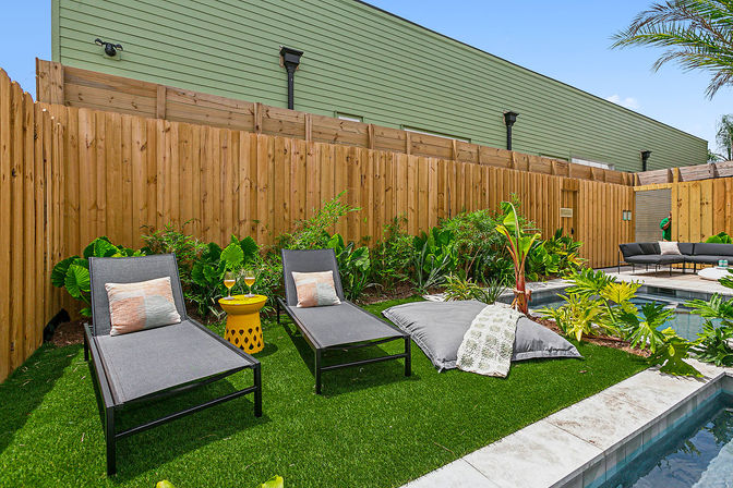 Residential backyard poolside lounge with two gray chaise lounges, yellow side table with drinks, oversized floor cushion on artificial turf, tropical plants and wooden privacy fence.