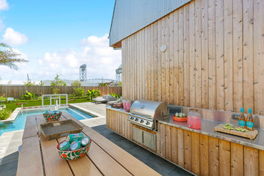 Sunny backyard patio with wooden outdoor kitchen and stainless-steel grill beside a rectangular pool, picnic table with bowls of canned drinks and a pitcher, lounge seating and a bridge in the distance.
