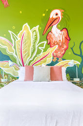 Bright tropical bedroom with a white bed, coral and beige pillows, twin bedside lamps, and a vibrant lime-green mural of oversized leaves and a stylized pink-orange bird.