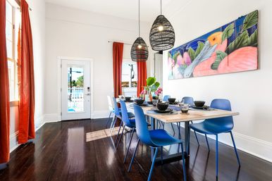 Bright coastal dining room with long wooden table set for eight, modern blue chairs, woven pendant lights, tropical wall mural, red curtains, glass door to a sunny patio/pool and dark hardwood floors.