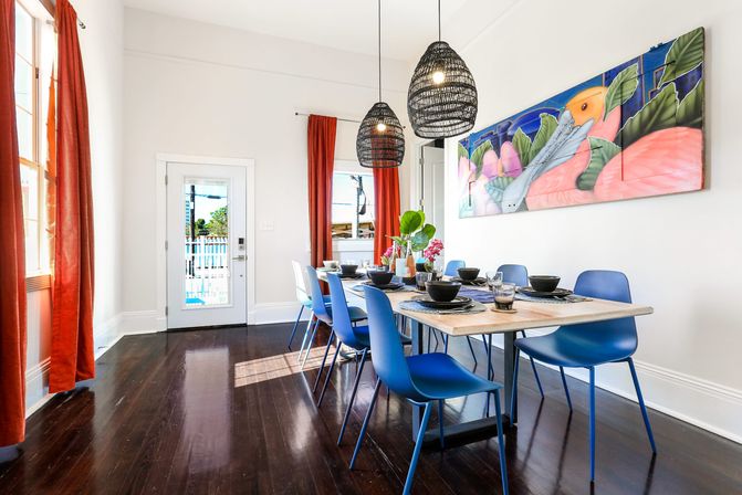 Bright coastal dining room with long wooden table set for eight, modern blue chairs, woven pendant lights, tropical wall mural, red curtains, glass door to a sunny patio/pool and dark hardwood floors.