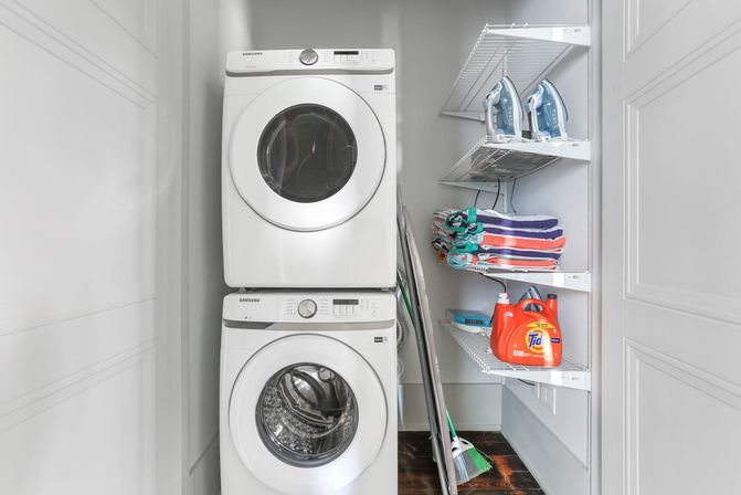 Compact laundry closet with stacked white front-load washer and dryer, wire shelves with folded colorful towels, three irons, orange detergent jug, broom and ironing board.
