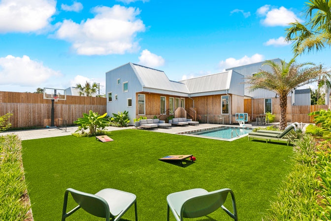 Sunny modern coastal backyard with sparkling pool, palm tree, artificial turf lawn with cornhole boards, outdoor lounge seating, basketball hoop, and metal-roofed contemporary house under a bright blue sky