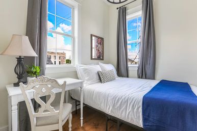 Sunny cozy urban bedroom with white linens and a blue throw, ornate white desk and chair with lamp and potted plant, two tall windows framed by gray curtains overlooking a neighboring brick building and blue sky.