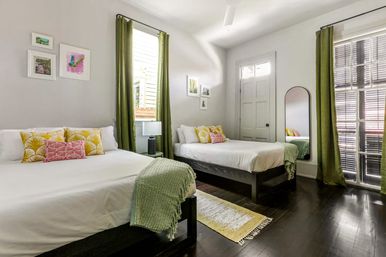 Sunlit modern guest bedroom with two white-made beds, green curtains and throws, yellow patterned pillows, hardwood floors, wall art and a tall mirror
