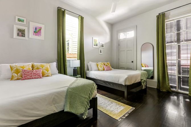 Sunlit modern guest bedroom with two white-made beds, green curtains and throws, yellow patterned pillows, hardwood floors, wall art and a tall mirror
