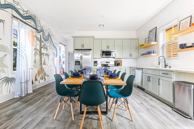 Bright contemporary open-plan kitchen and dining area with light gray shaker cabinets, stainless steel appliances, a wooden table set for eight with teal mid-century chairs, gray wood-look floors, white subway-tile backsplash, floating shelves, and a colorful mural on the side wall adding coastal-inspired flair.