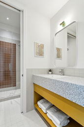 Contemporary apartment bathroom with terrazzo countertop, mustard-yellow vanity and open shelf of white towels, rectangular mirror and wall sconce, glass shower with brown square tiles and white tiled floor.