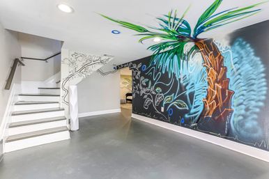 Finished basement with white staircase, gray floor, recessed ceiling lights and a vibrant palm-tree and leaf mural painted on a black accent wall