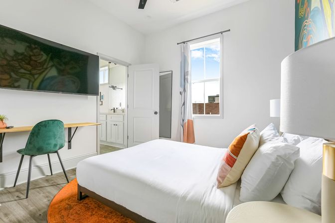 Bright modern bedroom with white walls, queen bed in crisp white linens and colorful throw pillows, orange round rug, green velvet desk chair at a floating wood desk, large wall-mounted TV, window with urban view and open door to a white-tiled ensuite bathroom.