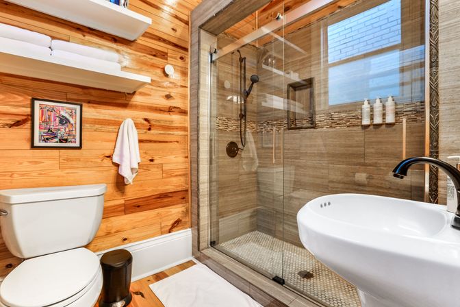 Cozy modern rustic bathroom with wood-plank walls, white toilet and pedestal sink, glass-enclosed tiled shower with built-in niche and floating shelves with towels.