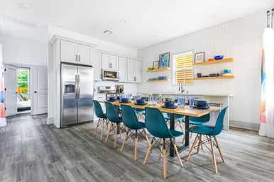 Sunlit modern open-plan kitchen and dining area with white cabinets, stainless steel refrigerator, floating shelves, long wood table set with blue dishes and teal chairs on gray wood-look flooring