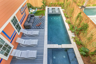 Aerial view of a narrow backyard oasis featuring a rectangular pool with attached spa, gray deck with three white lounge chairs, orange house with blue geometric trim, a cozy sofa and modern chairs on a patterned rug, small bistro table on grass, and a wooden fence planted with tropical greenery.