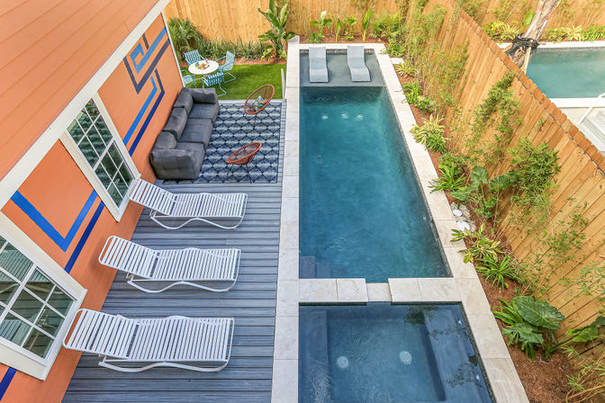 Aerial view of a narrow backyard oasis featuring a rectangular pool with attached spa, gray deck with three white lounge chairs, orange house with blue geometric trim, a cozy sofa and modern chairs on a patterned rug, small bistro table on grass, and a wooden fence planted with tropical greenery.