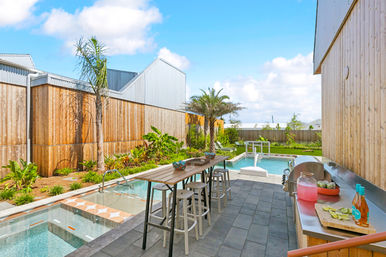 Sunlit modern backyard patio with plunge pool and spa, tall wooden privacy fence, tropical landscaping and palm trees, long outdoor bar with stools and a stainless-steel prep counter with drinks and sliced limes.