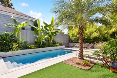 Sunny tropical backyard with a rectangular in-ground pool, palm tree and banana plants, lounge chairs on a concrete deck and green artificial turf