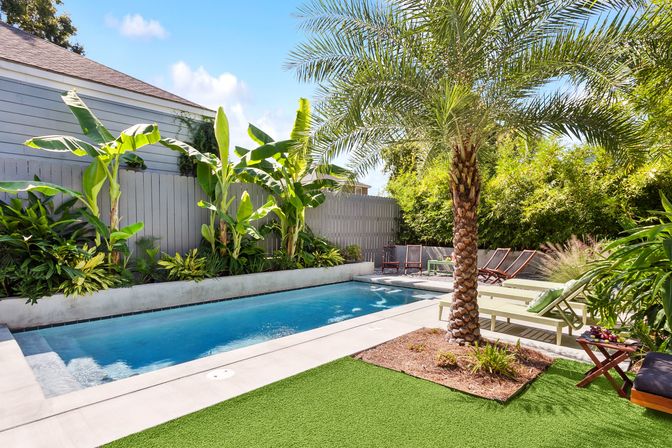 Sunny tropical backyard with a rectangular in-ground pool, palm tree and banana plants, lounge chairs on a concrete deck and green artificial turf